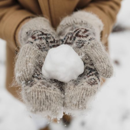 Hände in gemusterten Strickfäustlingen aus Wolle halten einen Schneeball im Schnee