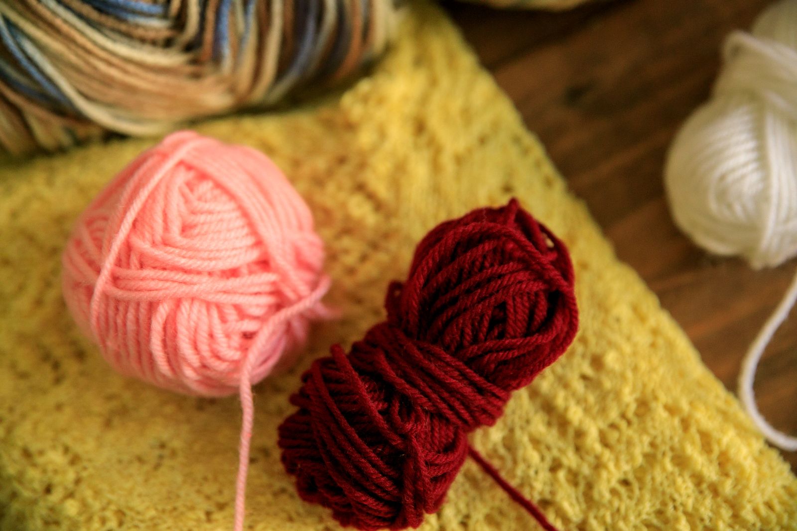 Nahaufnahme weicher Wollknäuel in Rosa, Bordeaux und Weiß auf einem gelben Strickstück