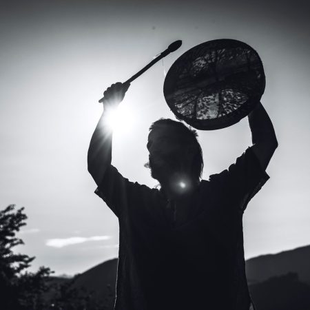Silhouette einer Person, die eine Rahmentrommel gegen die Abendsonne über den Kopf hält, dahinter Berge