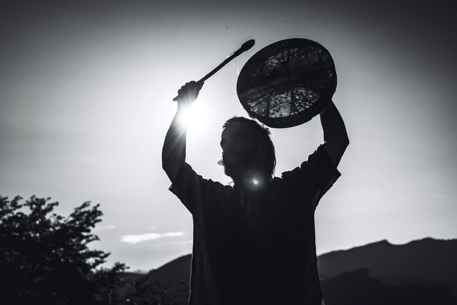 Silhouette einer Person, die eine Rahmentrommel gegen die Abendsonne über den Kopf hält, dahinter Berge